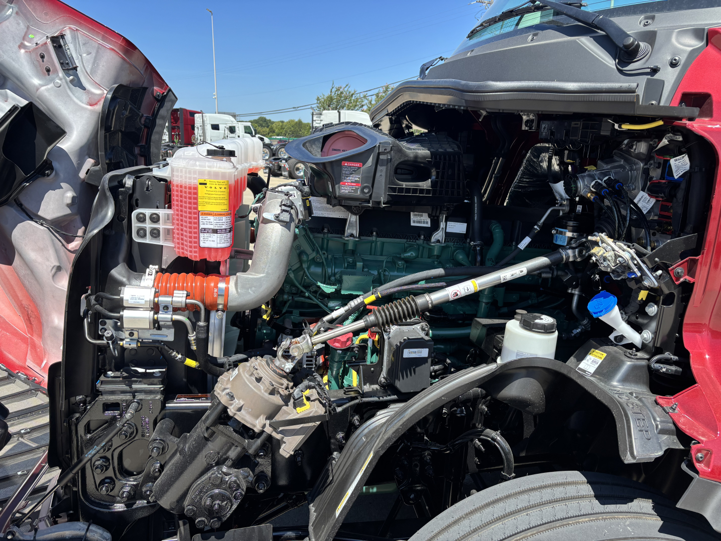 Volvo VNL64T860 engine bay showing diesel engine, coolant reservoir, and various hoses and wiring in a bright cab-over fold. Front left components visible.