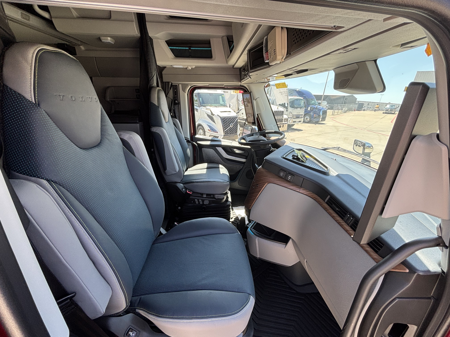 Interior of a Volvo VNL64T860 truck cab showing driver seat, steering wheel, dashboard, and front windshield with view of other trucks outside.