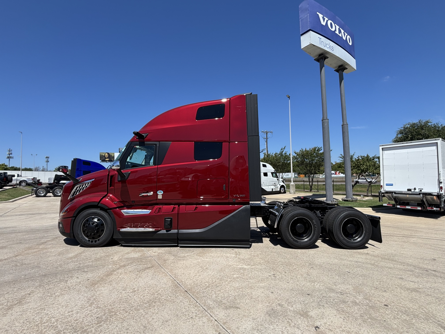 Volvo VNL64T860 truck tractor side profile in red with sleeper cab, showing chassis and rear axles, dealership lot in background.