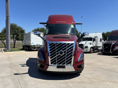 Front view of a red Volvo VNL64T860 semi truck with chrome grille and large bumper, parked in a lot among other trucks.