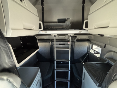Interior view of a new Volvo VNL64T860 sleeper cab showing upper bunk ladder, storage compartments, and seating area.