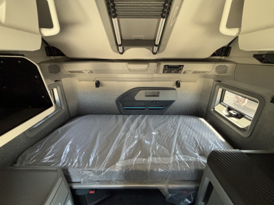 Truck cab interior of a 2027 Volvo VNL64T860 sleeper showing sleeping area with plastic-wrapped bed, overhead storage, window, and side panels.