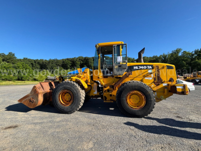 Hyundai HL740-7A wheel loader in profile with bucket attached, yellow with black accents, on gravel under clear blue sky.