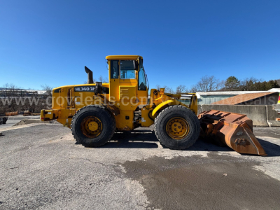 Wheel loader in yellow with front bucket on concrete, side view showing HL740-7 sticker and? visible branding. Clear blue sky.