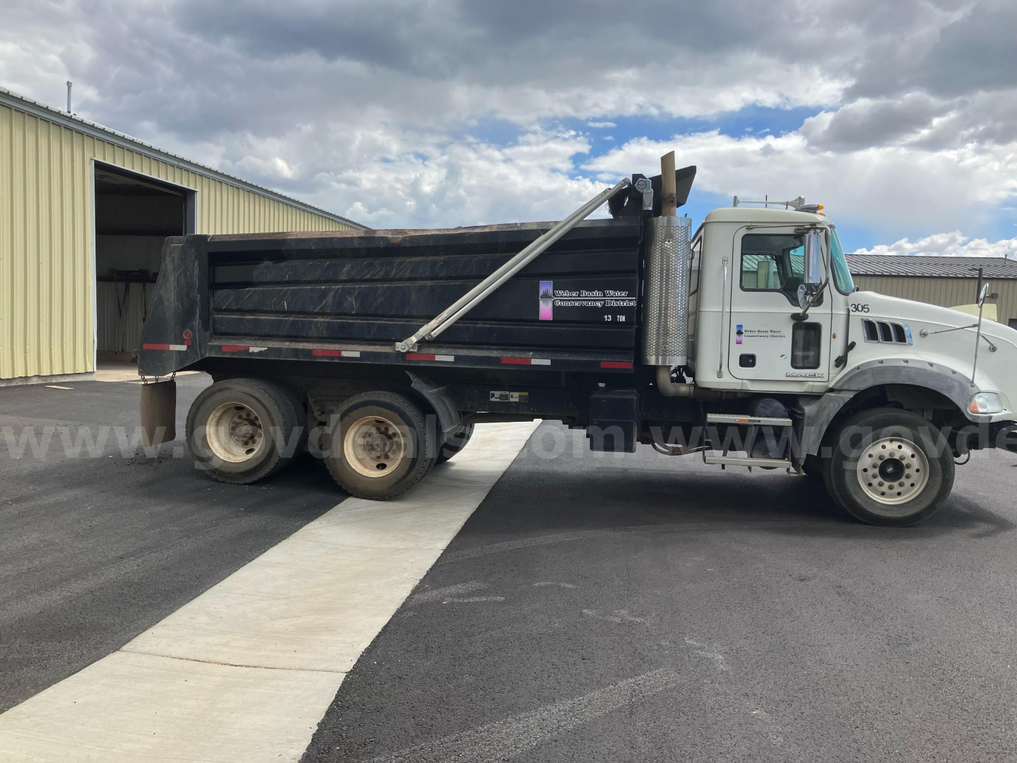 Dump truck side view showing Mack GU813 dump bed with hydraulic raise and dual rear axles, white cab, black dump body, on pavement.