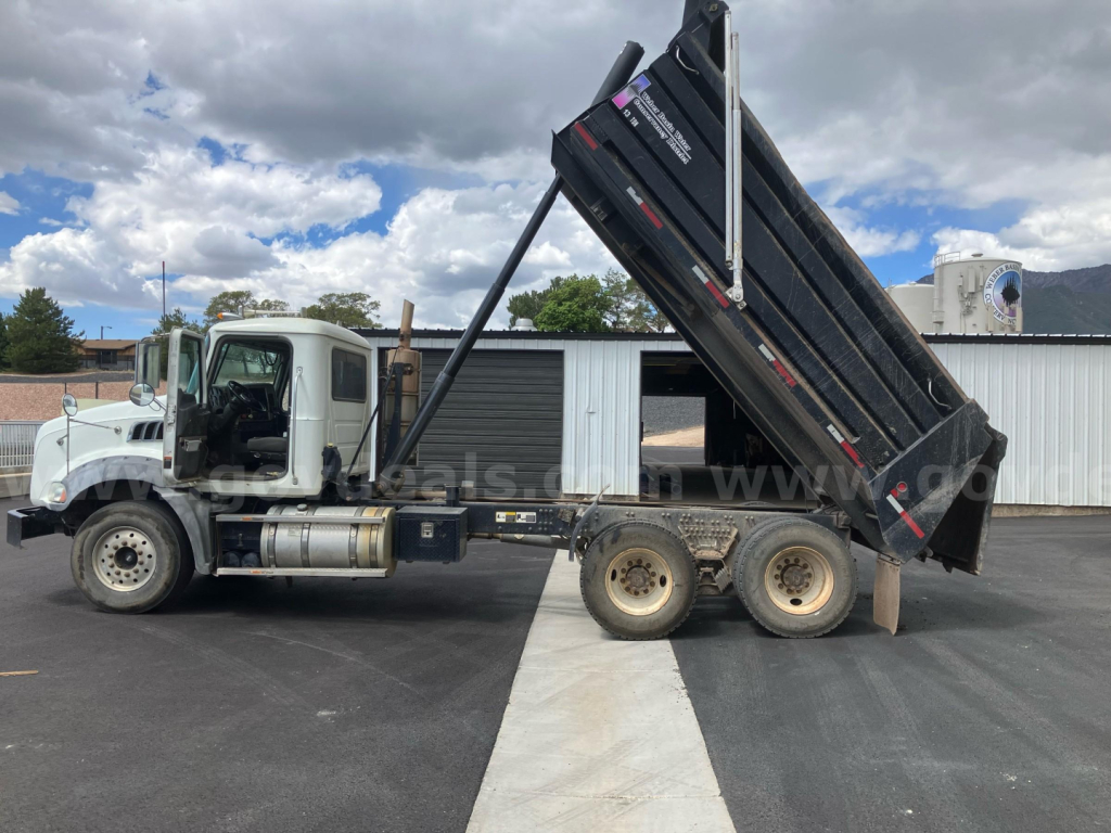 Dump truck with raised black dump bed on a Mack GU813 in-service condition, side view showing cab, dual rear axles, hydraulic raise mechanism, and loading area.