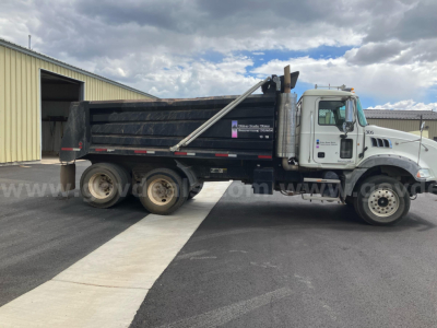 Dump truck side view showing Mack GU813 dump bed with hydraulic raise and dual rear axles, white cab, black dump body, on pavement.
