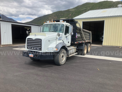 Mack GU813 dump truck in a parking lot showing front grille, cab, and dump bed with company decals; mountains in background.