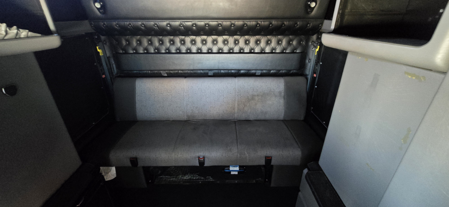 Kenworth W900 sleeper cab interior with black upholstered panels and storage compartments; visible are metal studs on the seatback and a gray bench seat.