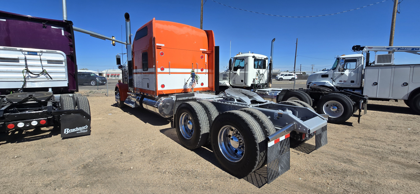 Kenworth W900 tractor chassis in a used trucks lot showing rear view with dual rear axles and orange sleeper cab in background.