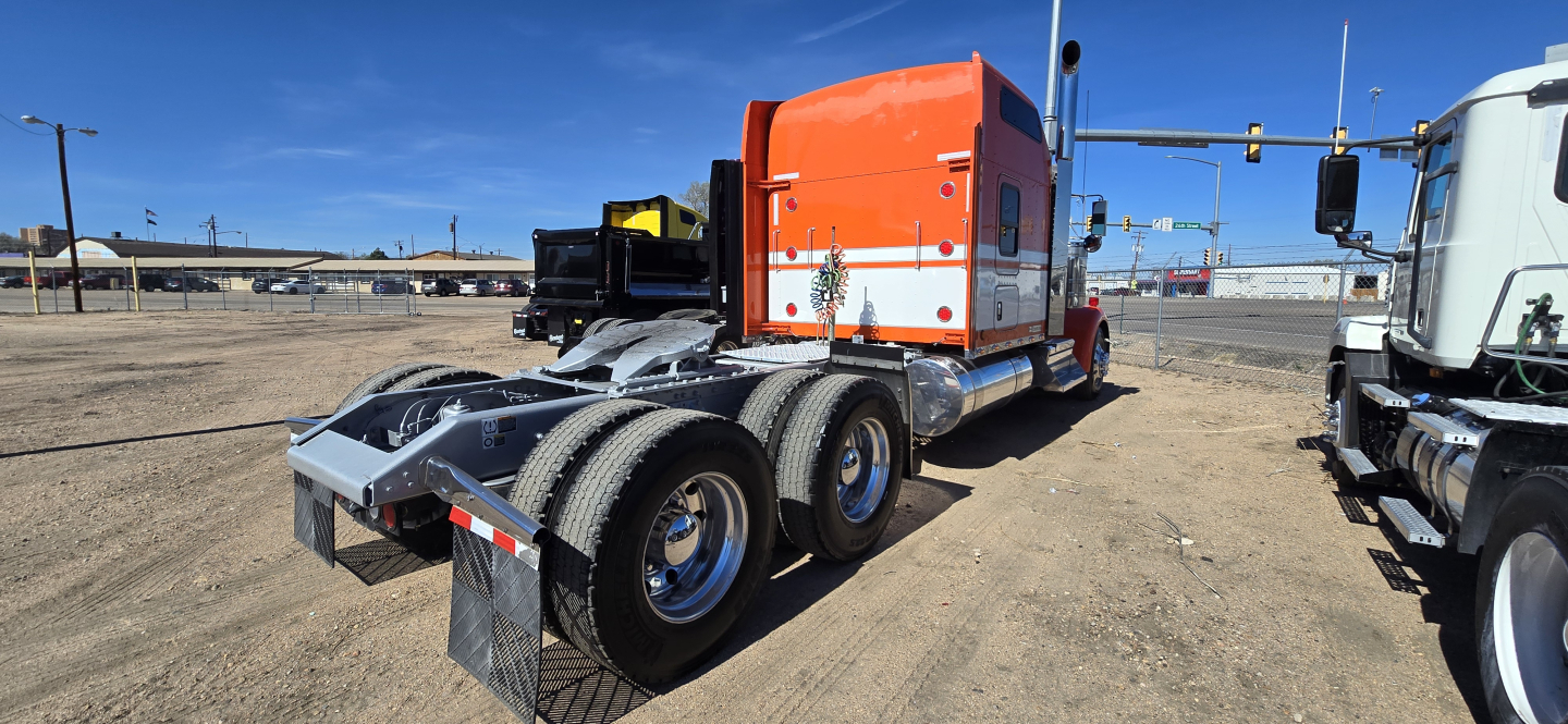 Kenworth W900 tractor in service view showing rear chassis and dual rear tires, orange sleeper cab with white stripe, chrome steps, and empty fifth wheel.