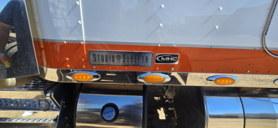 Kenworth W900 rear view showing Studio Sleeper badge and MHC emblem, with reflective chrome bumper and orange clearance/marker lights.