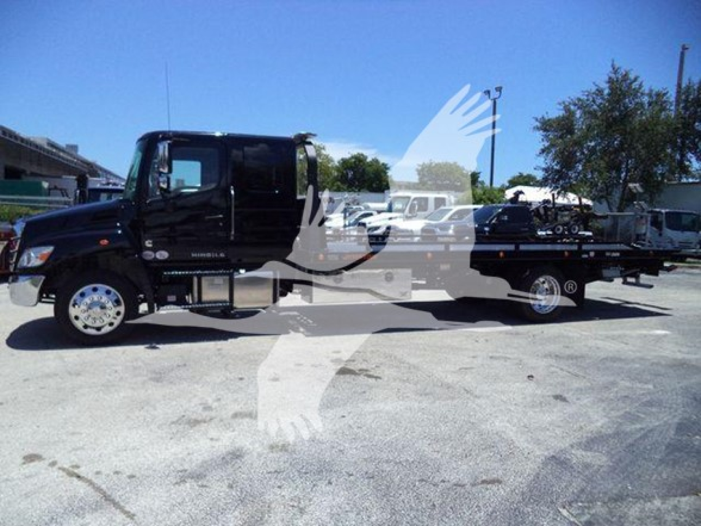 Black 2026 Hino L6 rollback tow truck in a lot with other trucks in background; side view shows cab and long bed with equipment. Text visible includes none specific on vehicle bodies.