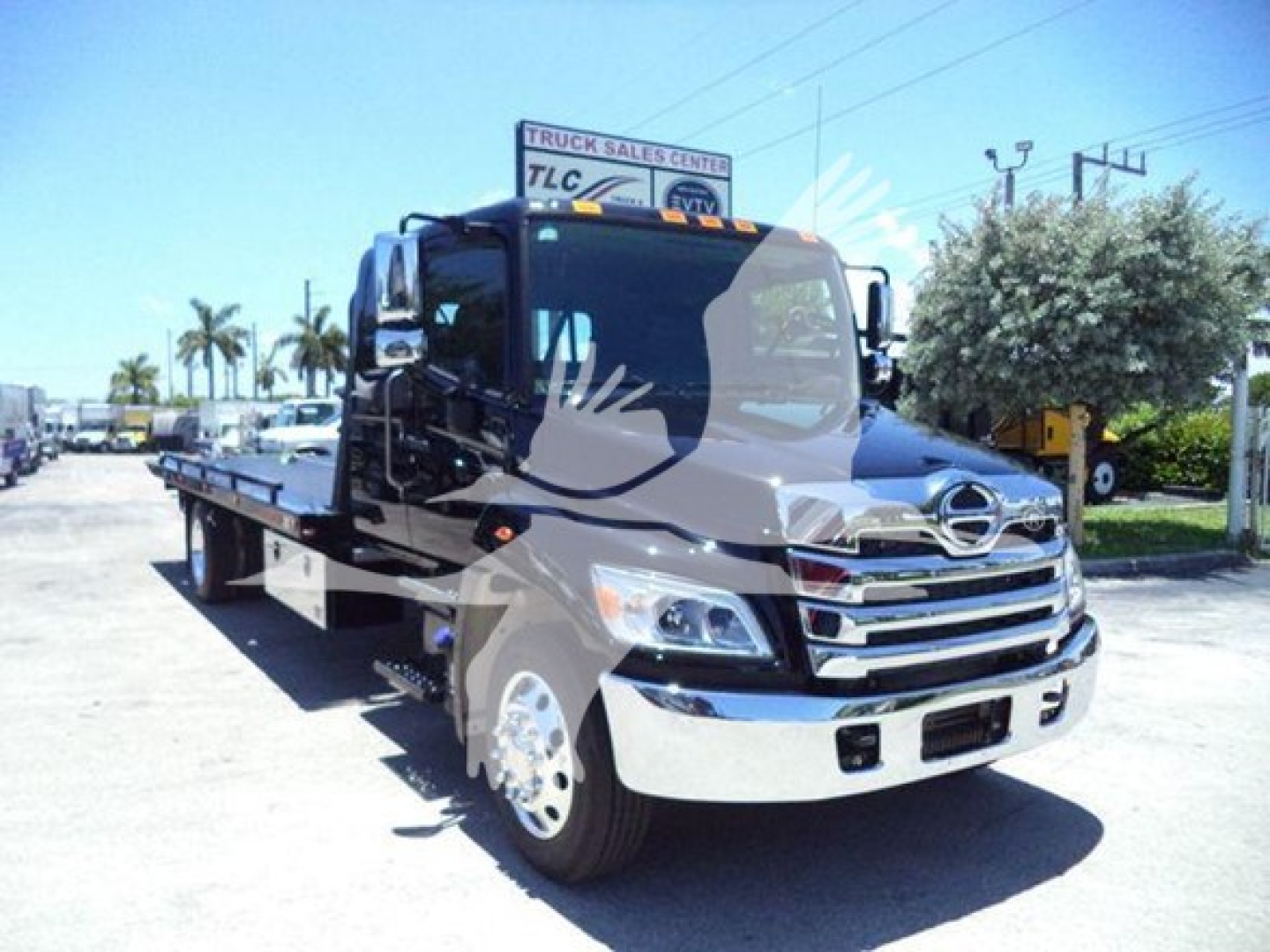 Flatbed tow truck in a dealership lot showing Hino L6 cab with chrome grille and side mirrors. Visible signage in background including TLC Truck Sales Center.
