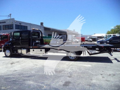 Flatbed tow truck side view showing rollback carrier on a 2026 Hino L6 with cab and chassis in black, dealership lot background.