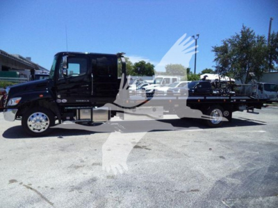 Black 2026 Hino L6 rollback tow truck in a lot with other trucks in background; side view shows cab and long bed with equipment. Text visible includes none specific on vehicle bodies.