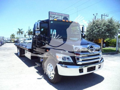 Flatbed tow truck in a dealership lot showing Hino L6 cab with chrome grille and side mirrors. Visible signage in background including TLC Truck Sales Center.