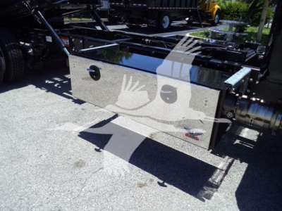 Truck bed toolbox with chrome reflective surface and logos; visible text includes small round button-like handles and a badge showing 'Chevrolet' or related emblem, with a dark cylindrical exhaust/ladder area; shadows of hands overlay.
