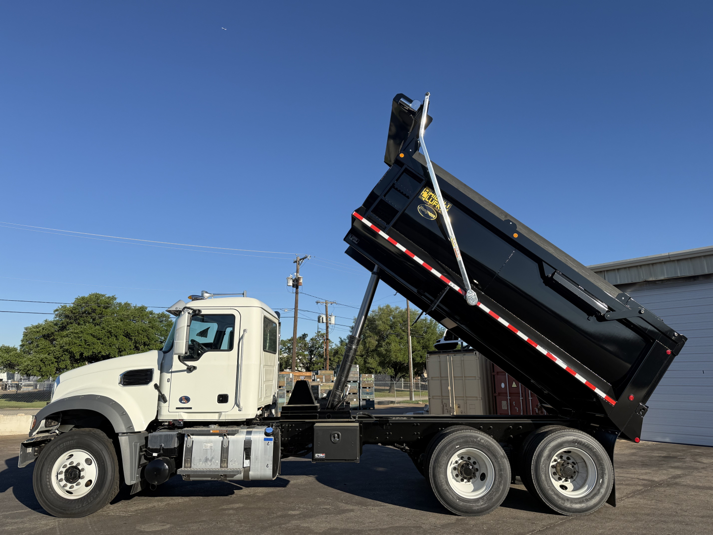 Front-left side view of a 2026 Mack GR64F dump truck with its black dumping bed raised, white cab, three rear axles, chrome fuel tanks, and hydraulic mechanism visible.