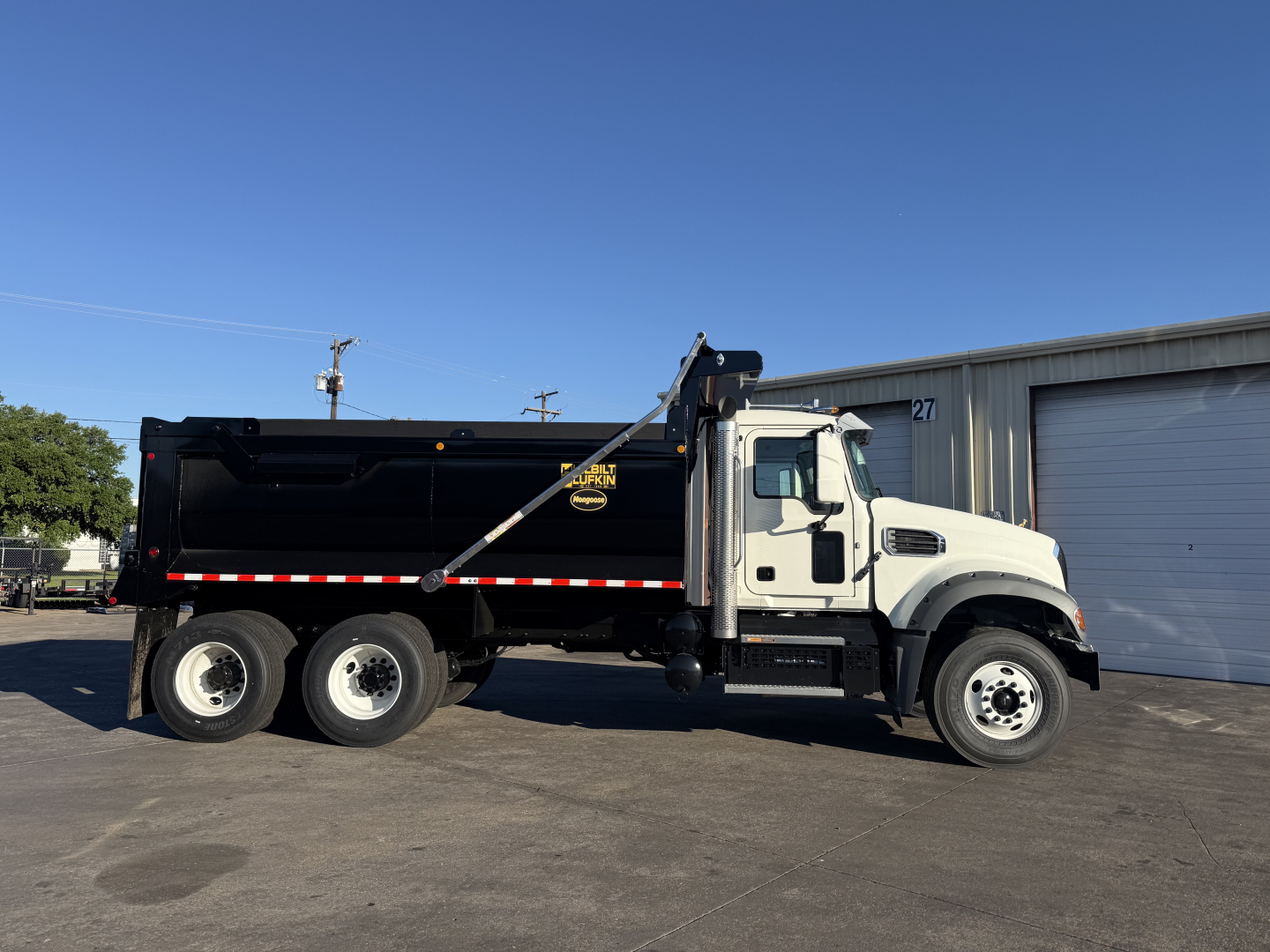 Dump truck side view showing a black Mack GR64F dump bed with white cab, three axles, and hydraulic dump mechanism.