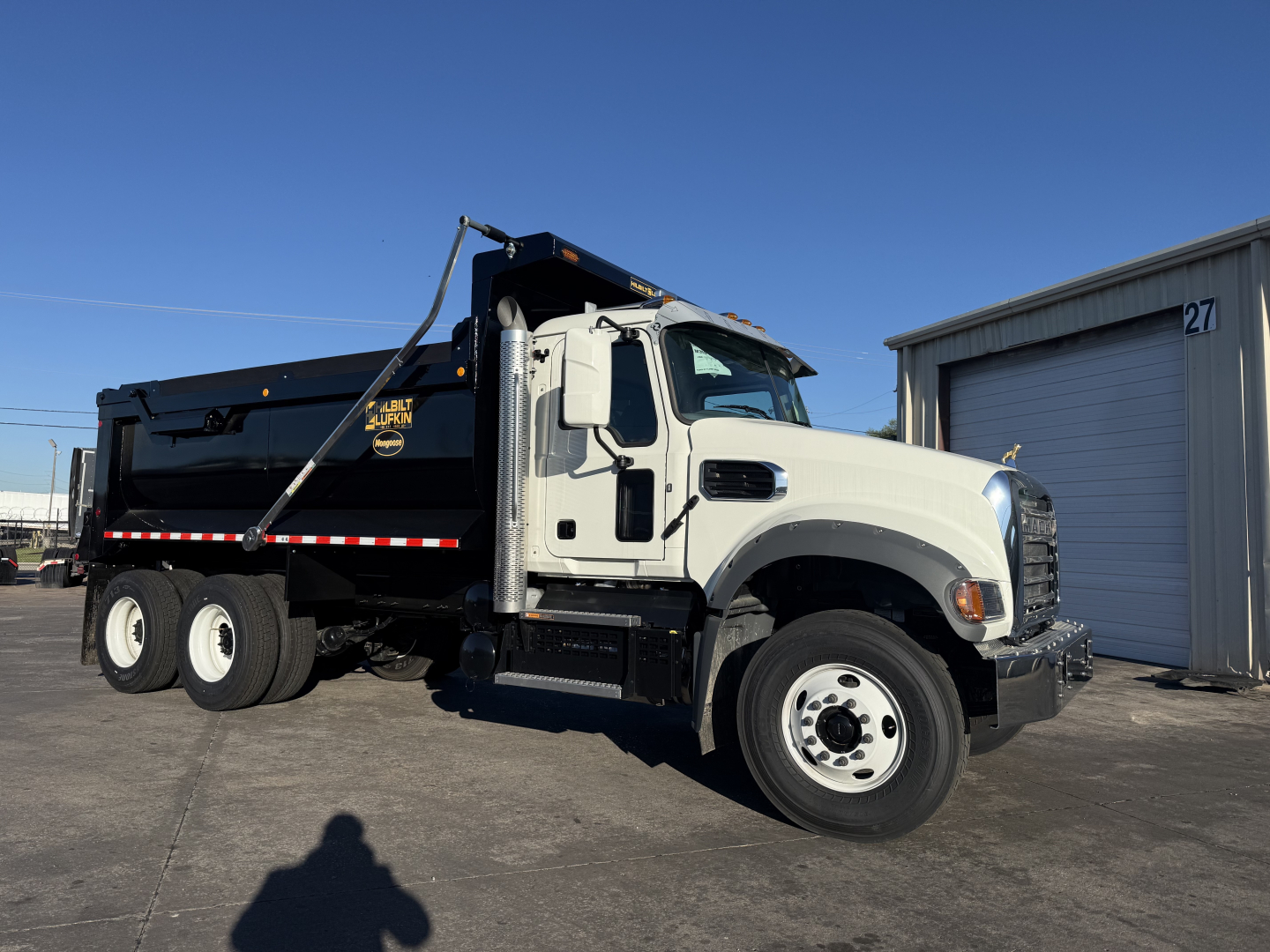 Dump truck side view showing Mack forward cab with white cab and black dump body, chrome exhaust stack, hydraulic arm, and reflective stripes. Visible text on cab door reads 'MACK' and on the dump body possibly logos, but not legible.