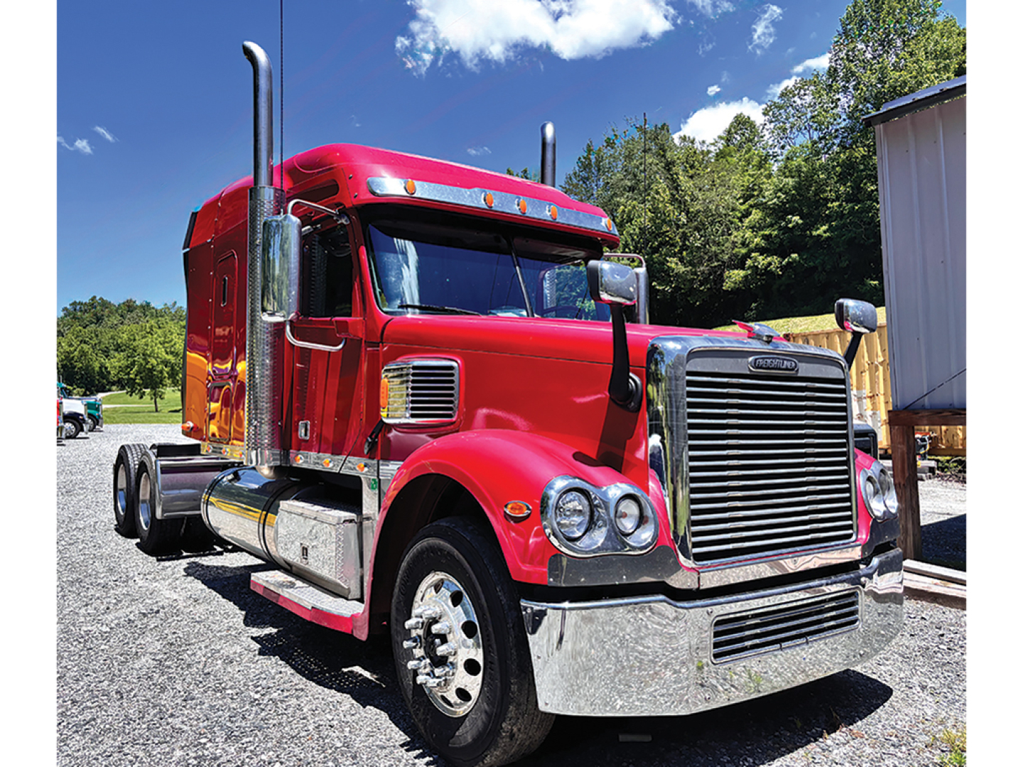 Red Freightliner Coronado tractor truck in a lot showing large grille, dual exhaust stacks, chrome bumper, and side fuel tanks.