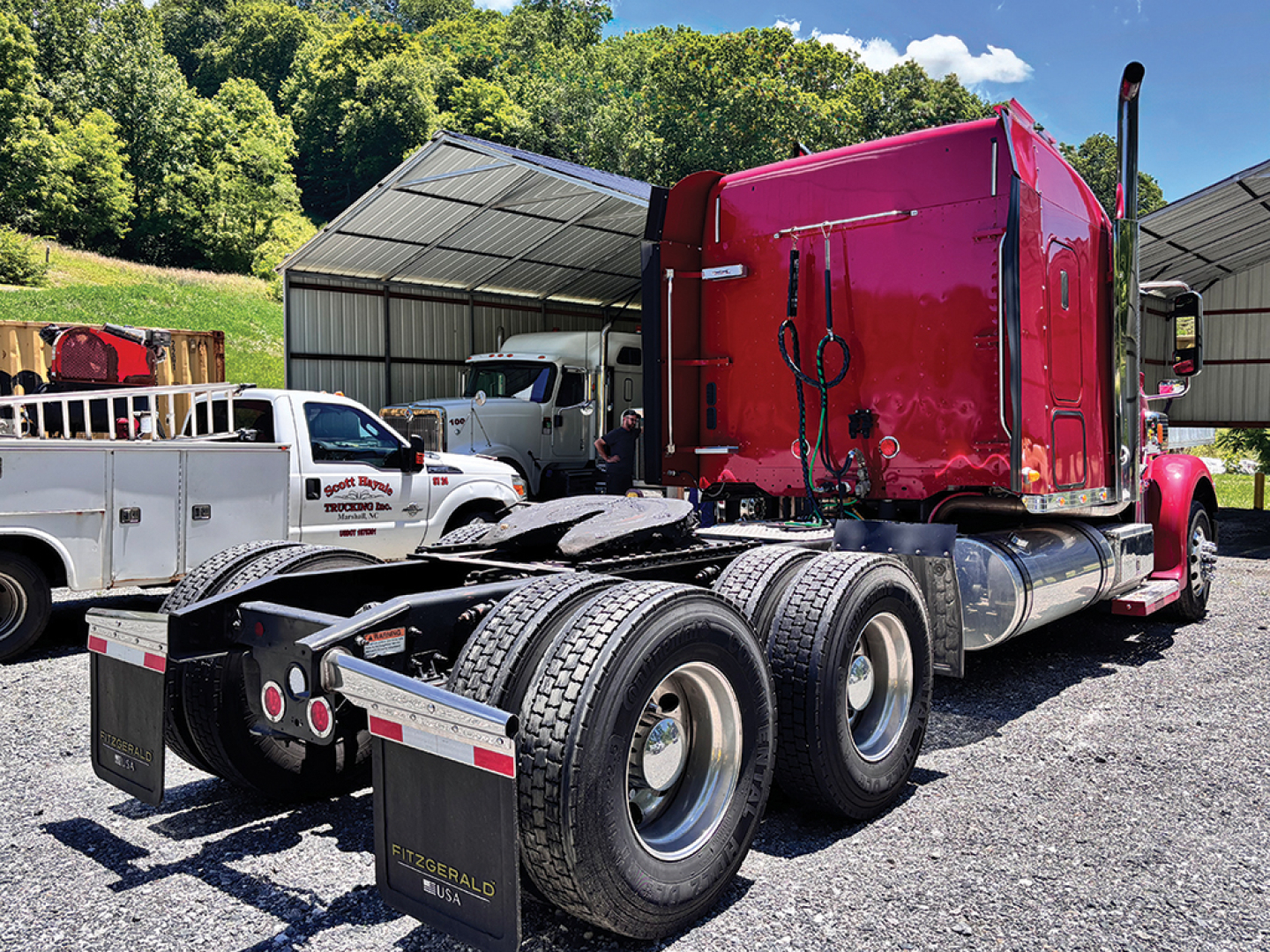 Truck chassis with red Freightliner Coronado sleeper cab, dual rear axles, fifth wheel, and tires; side mirrors and fuel tanks visible; equipment yard background.