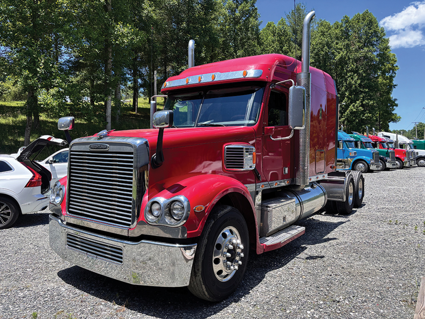 Freightliner Coronado truck in red, front cab view with chrome grill, large exhaust stacks, and dual rear axles. Vehicle shows stock number and generalized drivetrain area.