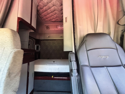 Cab interior of a Freightliner Coronado tractor showing sleeper area and driver seat with Legacy upholstery; compact rear cargo area visible with steps and cabinetry.
