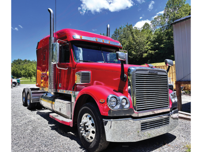 Red Freightliner Coronado tractor truck in a lot showing large grille, dual exhaust stacks, chrome bumper, and side fuel tanks.
