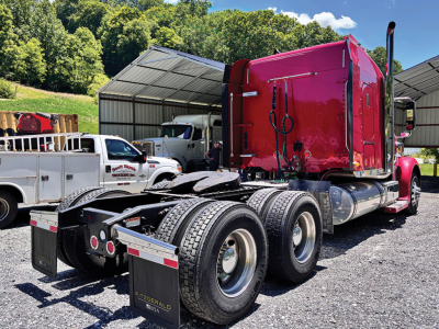 Truck chassis with red Freightliner Coronado sleeper cab, dual rear axles, fifth wheel, and tires; side mirrors and fuel tanks visible; equipment yard background.