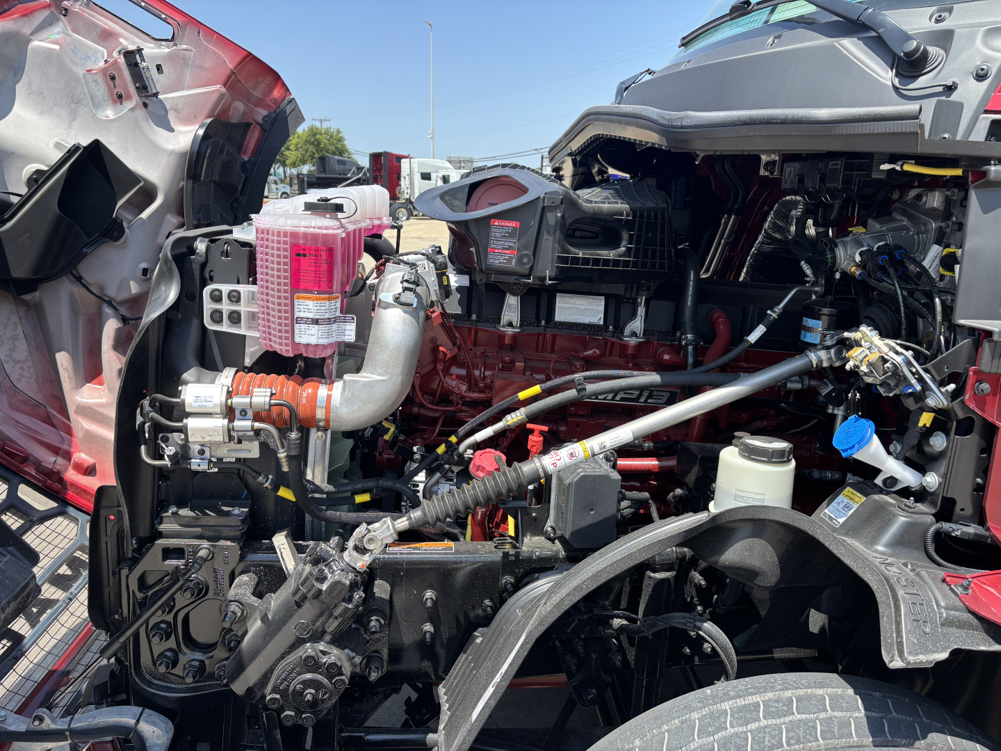 Engine bay of a 2027 Mack PR64T76H truck showing coolant reservoir, air intake, hoses, and wiring harnesses visible under open hood.