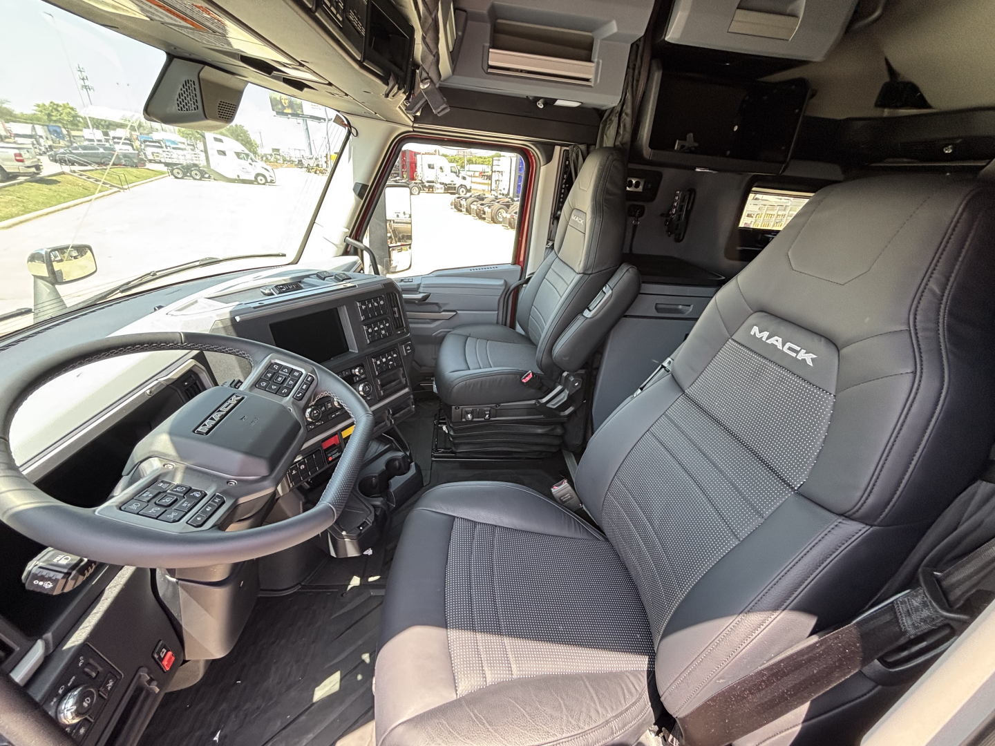 Interior of a 2027 Mack PR64T76H truck cab showing driver seat and passenger seat with Mack logo, dashboard, steering wheel, and overhead storage.