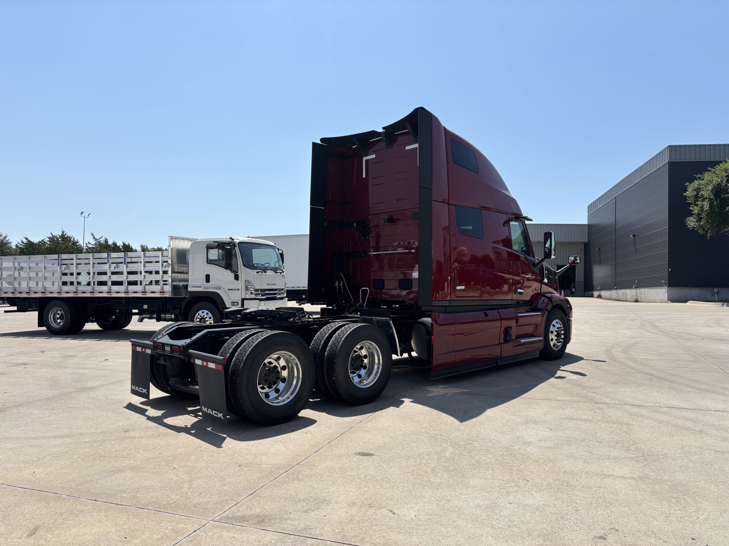 Truck cab and chassis of a new 2027 Mack PR64T76H tractor, red with sleeper, rear triple axle, side view showing long hood and elevated sleeper cab.