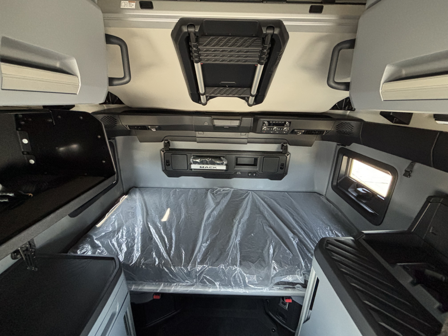 In-cab view of a new Mack PR64T76H truck sleeper showing overhead storage, bed area with plastic covering, and compact storage compartments.