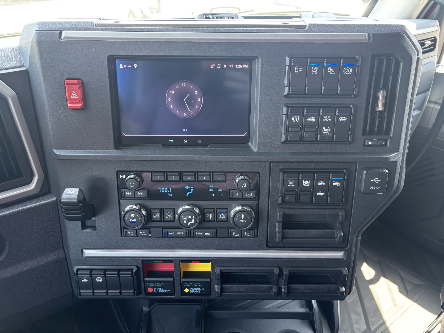 Mack PR64T76H truck dashboard showing touchscreen display, climate controls, and various switches; center console with storage slots and colored hazard/auxiliary buttons.