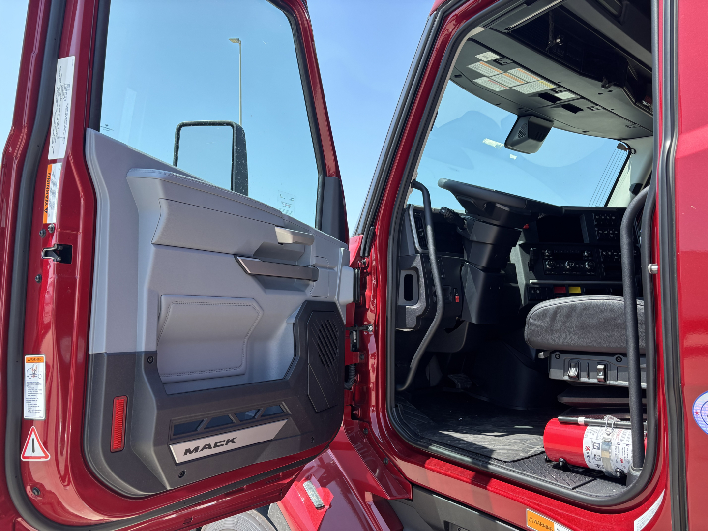 Truck cab interior and door panel of a red 2027 Mack PR64T76H with open driver door, showing dashboard, steering wheel, and seat; visible Makc badge on door sill.