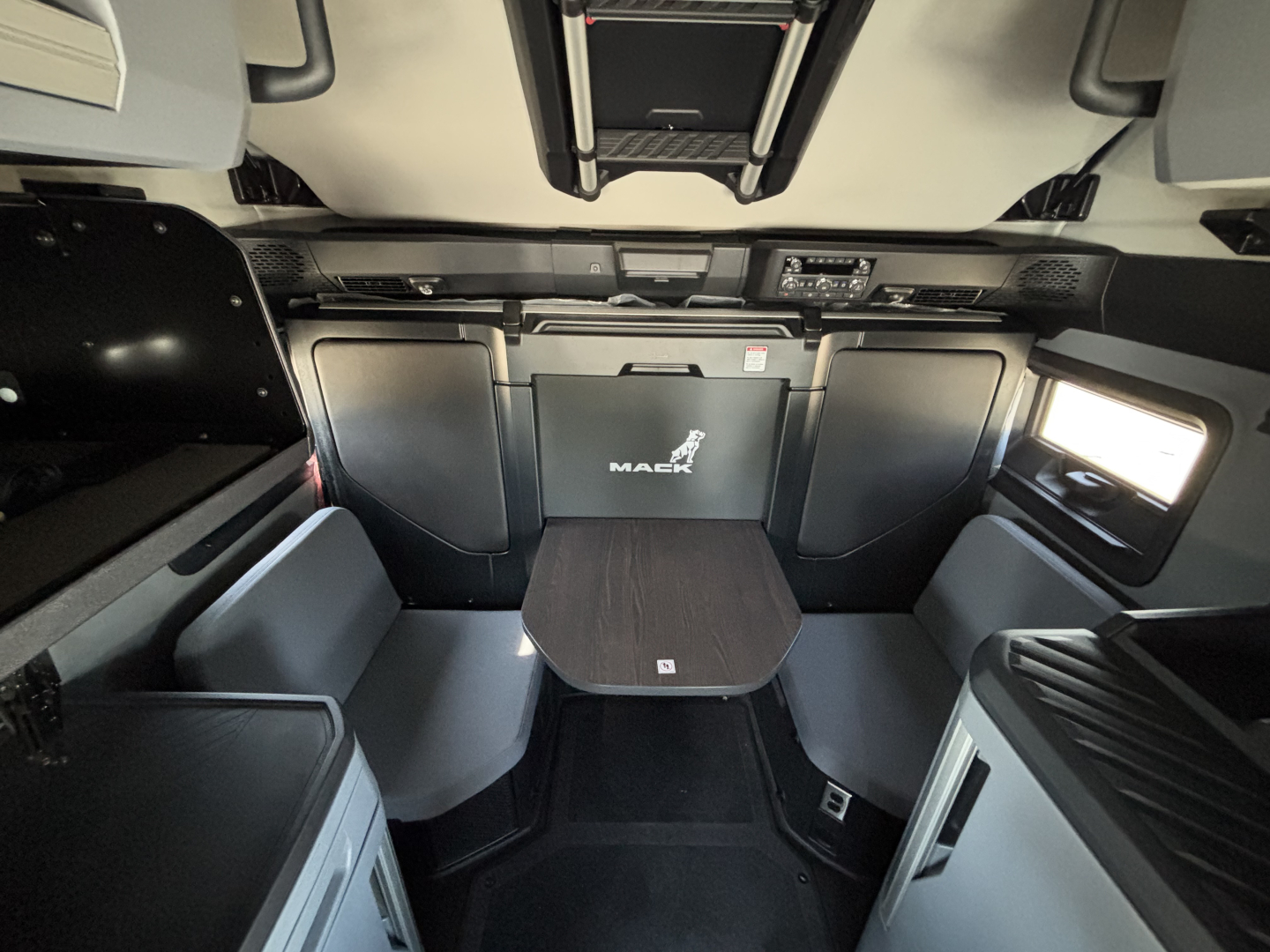 Mack PR64T76H truck interior showing sleeper cab with seating area and table, black and gray theme, overhead storage, window on right.