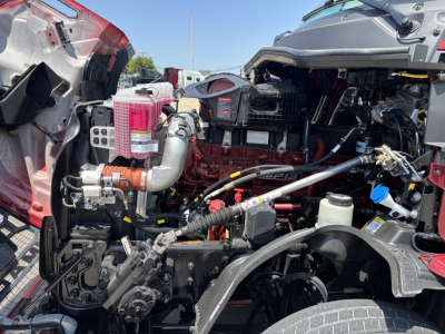 Engine bay of a 2027 Mack PR64T76H truck showing coolant reservoir, air intake, hoses, and wiring harnesses visible under open hood.