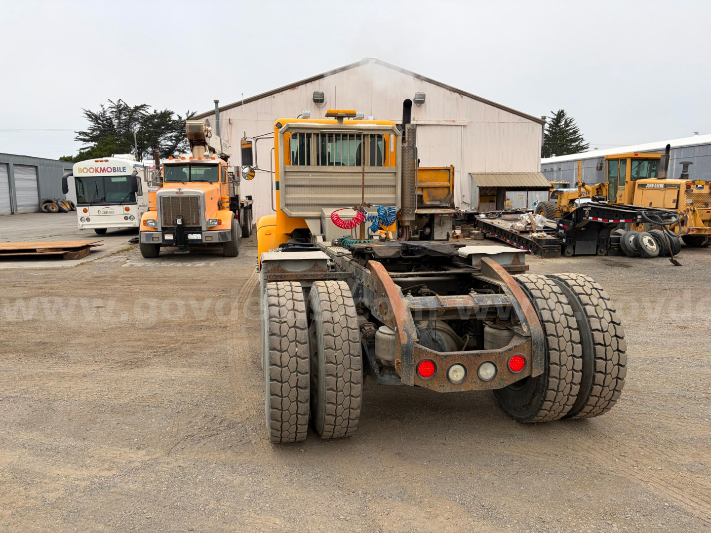 Peterbilt 367 truck chassis rear view with dual rear axles and yellow cab, other trucks and a warehouse in the background.