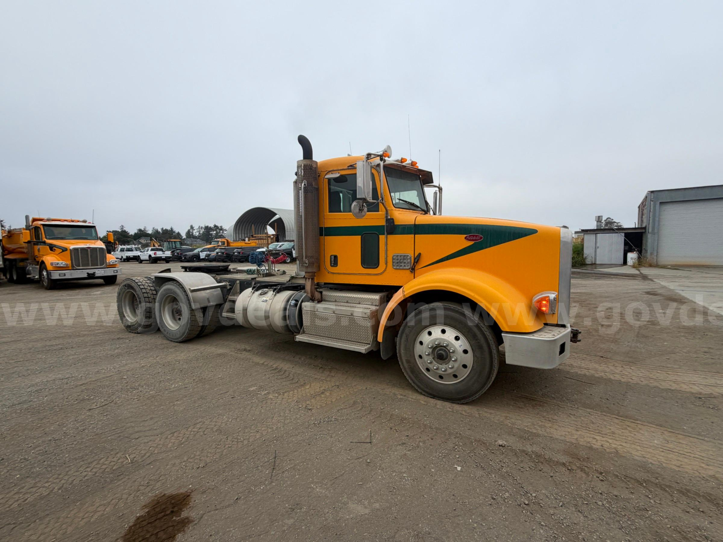 Peterbilt 367 cab-forward tractor in a gravel lot, yellow with green stripe, side view showing fuel tanks and chassis.