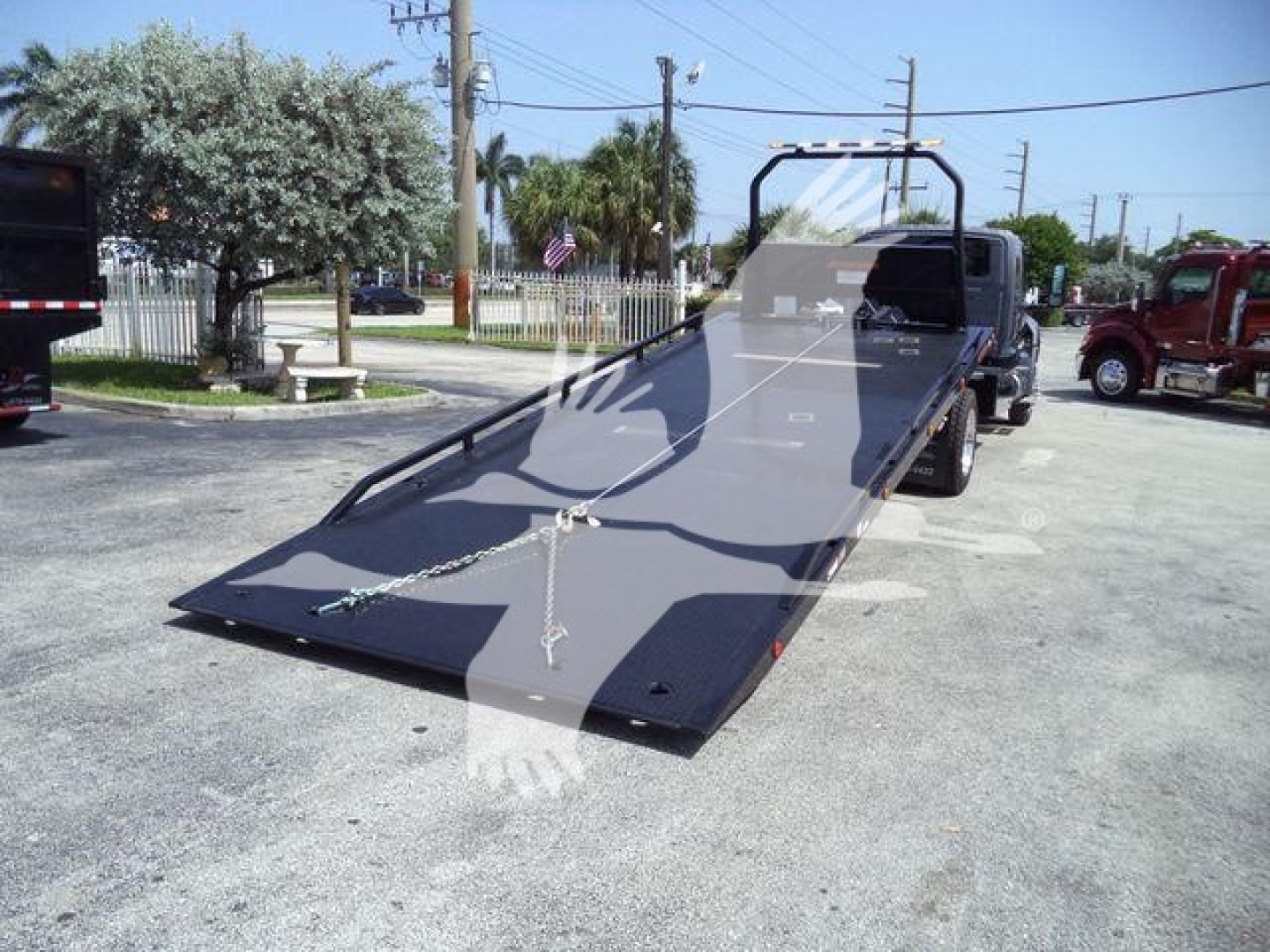 Flatbed tow truck with a long gray ramp on a rollback bed. Visible text includes no clear brand name but shows a chain and hook near the ramp. Background shows other trucks and a fenced area.