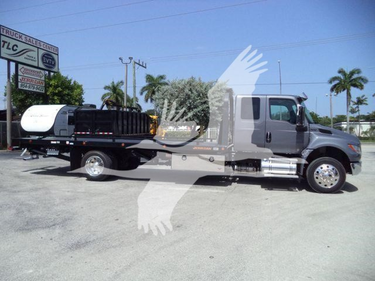 Truck with a flatbed rollback trailer and a white cylindrical tank attached, side view showing cab and dual rear wheels, International MV model branding not visible.