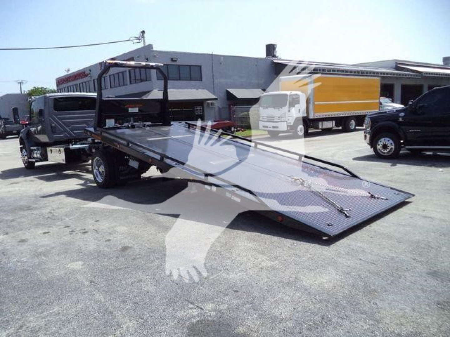 Truck tow rollback equipment with flatbed ramp and hitch, exterior lot view with other trucks in background.