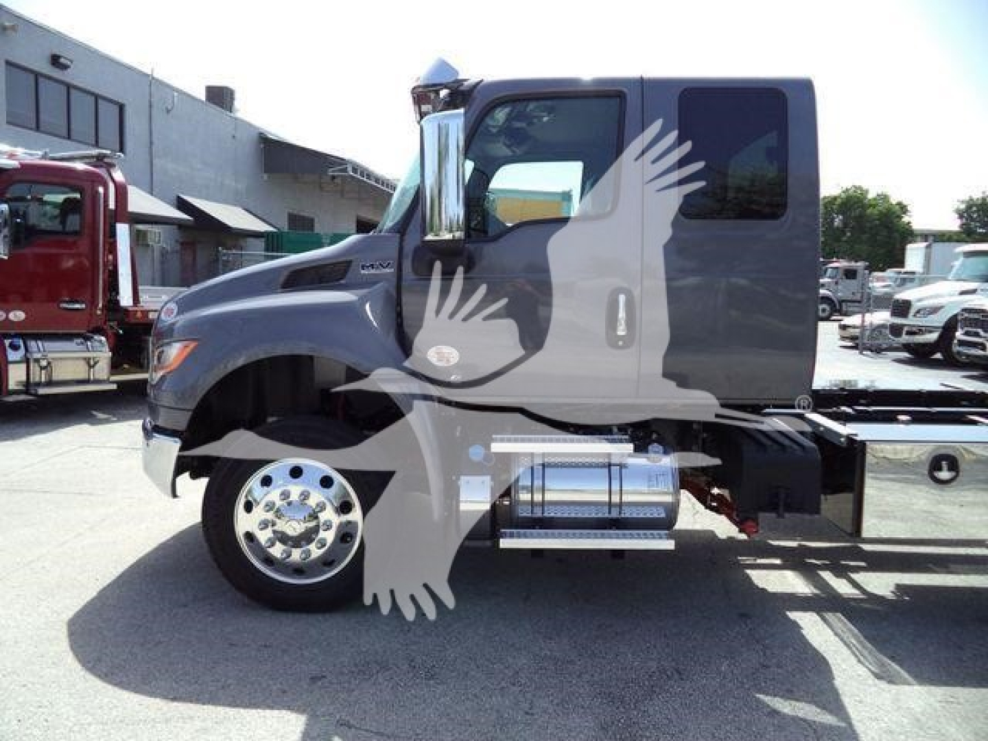 Truck side view showing a gray International MV cab with chrome fuel tank and steps, rollback bed visible, wheel front tire, side window, and dealership lot background.