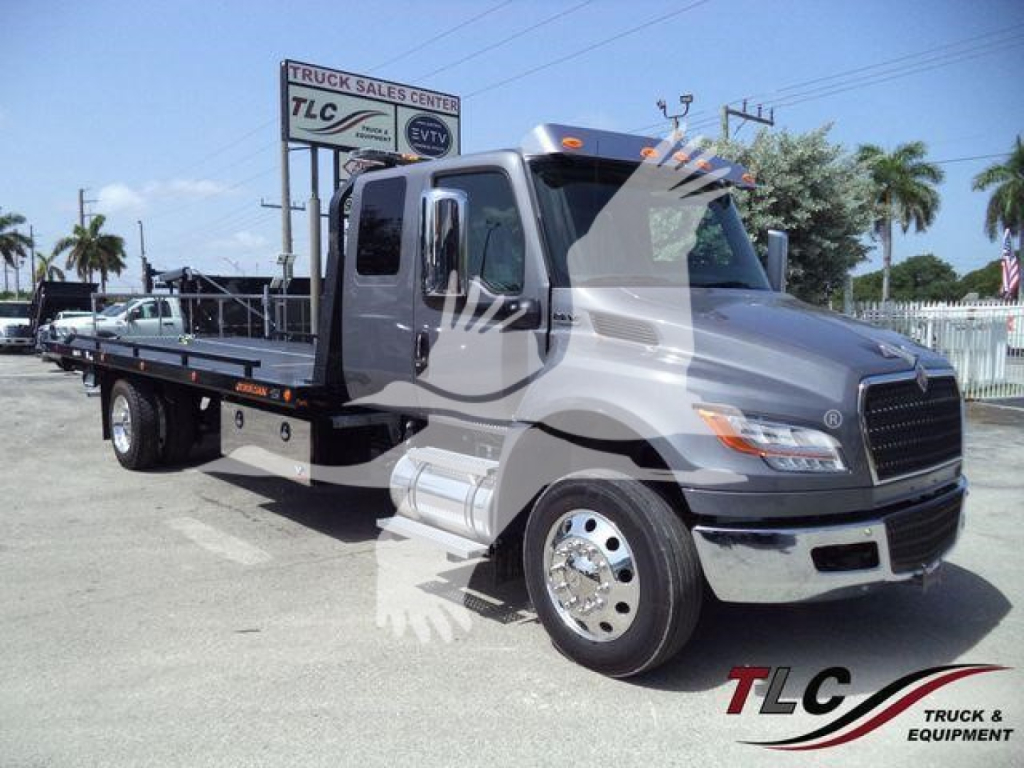 Flatbed tow truck from side/front view with cab and long flat deck, MV model plate visible.