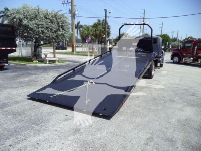 Flatbed tow truck with a long gray ramp on a rollback bed. Visible text includes no clear brand name but shows a chain and hook near the ramp. Background shows other trucks and a fenced area.