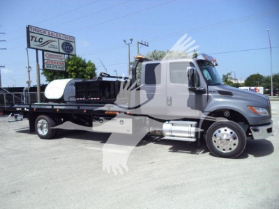 International MV rollback truck with a flatbed and a white cab; visible text includes 'TRUCK SALES CENTER TLC' on a sign in background; trailer bed shows some equipment and a logo on the side.