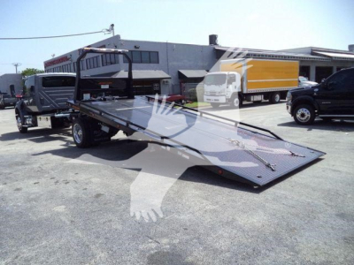 Truck tow rollback equipment with flatbed ramp and hitch, exterior lot view with other trucks in background.