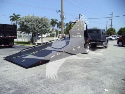 Truck with a large flatbed ramp in a rollback tow setup, black cargo bed and towing vehicle, pickup trucks in background, outdoor lot, clear weather.